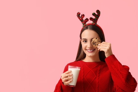 Young woman in Christmas reindeer horns with milk and cookies on pink backgroundの写真素材