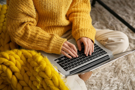 Beautiful young woman with warm yellow plaid sitting on floor and using laptop in living room....