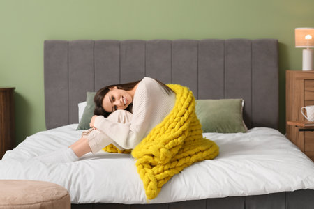 Beautiful young woman with warm yellow plaid sitting on bed in bedroomの写真素材