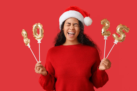 Happy African-American woman in Santa hat and with figure 2025 made of balloons on red background....
