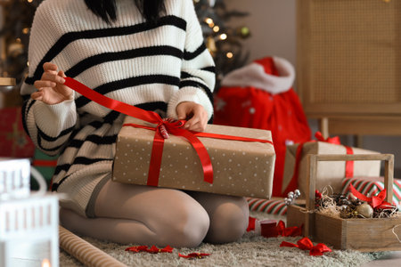 Beautiful young woman with decor tying bow while wrapping Christmas gift box at home, closeupの写真素材