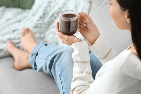 Beautiful young woman with glass cup of hot cocoa sitting on sofa at home. Christmas celebrationの写真素材