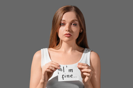 Crying young woman holding paper with text I'M FINE on dark background