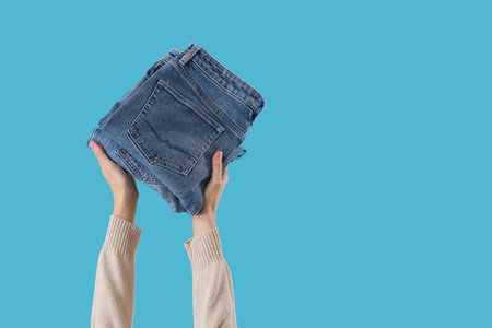 Female hands with stack of stylish jeans on blue background, closeup