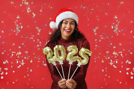 Happy African-American woman in Santa hat and with figure 2025 made of balloons on red background....