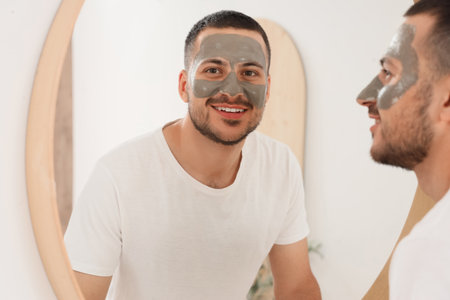 Handsome young happy man with cosmetic clay mask on face near mirror in bathroom at homeの写真素材