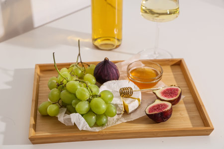 Wooden board with sweet ripe grapes, figs, camembert cheese, honey and bottle of wine on white...