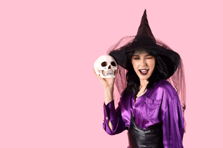 Beautiful young happy woman dressed as witch for Halloween with human skull on pink backgroundの写真素材