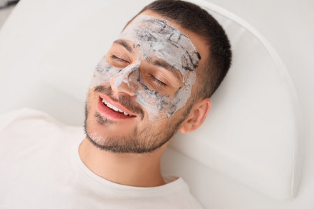 Handsome young happy man with cosmetic bubble mask on face lying in spa salonの写真素材