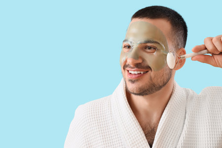 Handsome young happy man in bathrobe with cosmetic gel mask on face doing massage against blue backgroundの写真素材