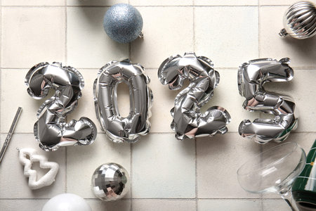 Figure 2025 made of foil balloons with Christmas decorations and glass on white tile background