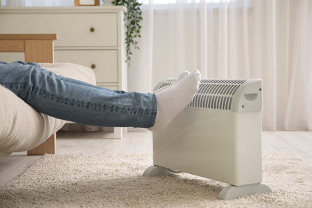 Young man sitting on sofa near electric heater in living room, closeup