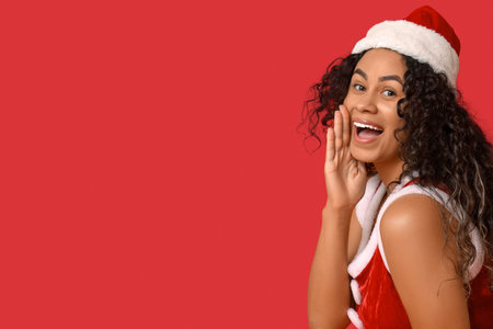 Beautiful young happy African-American woman in sexy Santa costume on red backgroundの写真素材