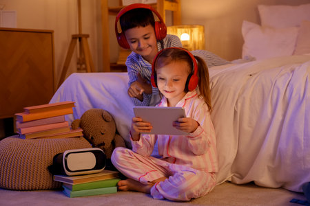 Cute happy little kids with tablet, headphones and books playing video games on floor in evening at homeの写真素材