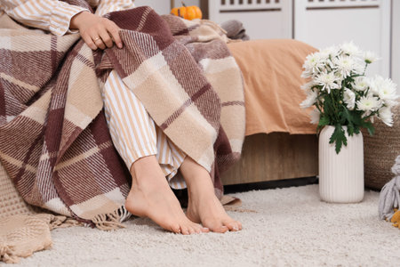 Beautiful young woman with warm plaids and vase of flowers sitting in bedroom at home, closeup