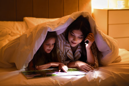 Happy mother and her little daughter with flashlight reading fairy-tale under blanket in bed at...