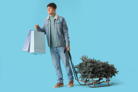 Young man with shopping bags and Christmas tree on sledge against blue backgroundの写真素材