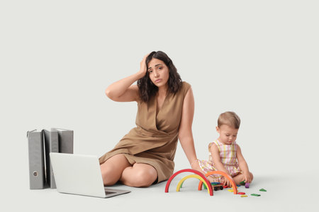 Tired businesswoman and her little baby playing with toys on light backgroundの写真素材