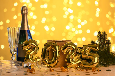 Bottle of champagne with glasses, Christmas gift box and figure 2025 made of balloons on table against blurred lightsの写真素材
