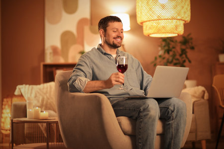 Handsome man with glass of wine and laptop having online date at home in eveningの写真素材