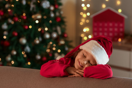 Cute happy girl in Santa hat near sofa on Christmas eve at homeの写真素材