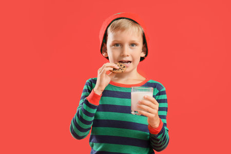 Cute happy little boy in Christmas pajamas with cookies and glass of milk on red backgroundの写真素材
