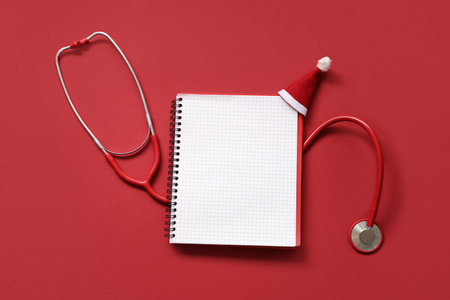 Stethoscope with notebook and decorative Christmas hat on red backgroundの写真素材