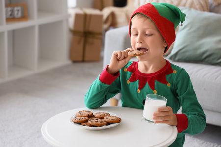 Cute little boy dressed as Christmas elf eating cookies with cup of milk at homeの写真素材