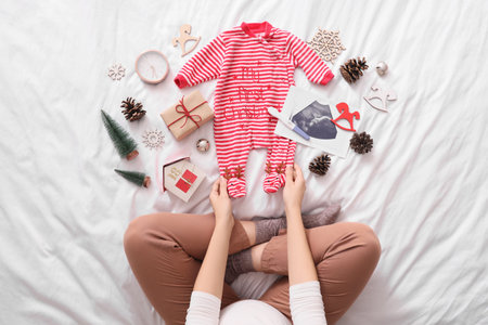 Young pregnant woman with pregnancy test, baby clothes, sonogram image and Christmas decorations sitting on bed, top viewの写真素材