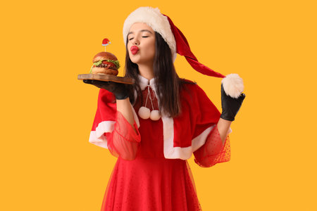 Pretty young woman in Santa costume with tasty burger blowing kiss on yellow backgroundの写真素材