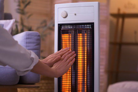 Young woman warming hands near electric heater in evening at home, closeupの写真素材