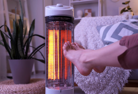 Young woman warming legs near electric heater in evening at home, closeupの写真素材