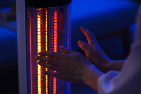 Young woman warming hands near electric heater in evening at home, closeup