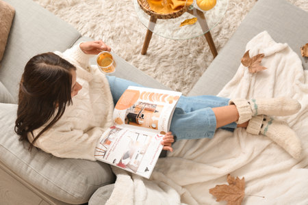 Beautiful young woman with cup of green tea and magazine on autumn day at homeの写真素材