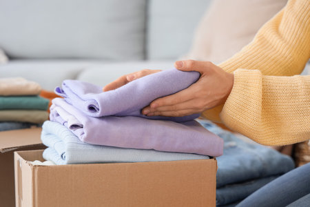Young woman putting stack of clean clothes into cardboard box at home, closeup