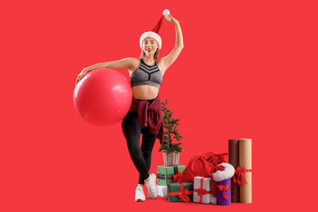 Sporty young woman with fitball and Christmas gift boxes on red backgroundの写真素材