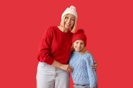 Beautiful woman with her cute daughter in sweaters and hats on red stylish backgroundの写真素材