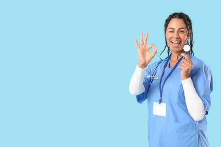 Young African-American female doctor with stethoscope and badge showing OK gesture on blue backgroundの写真素材