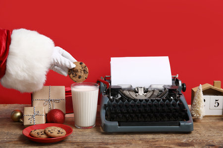 Santa Claus hand with Christmas gift boxes, cookies, glass of milk and typewriter at wooden tableの写真素材