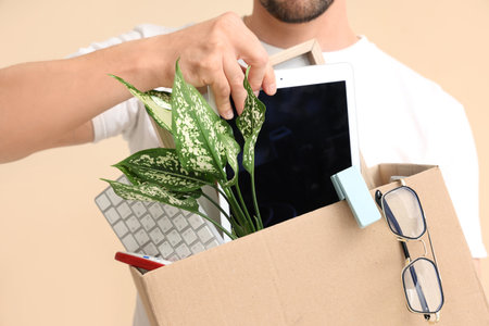 Fired young man packing his personal stuff in box on beige background, closeup