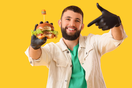 Young man in black gloves pointing at tasty burger on yellow backgroundの写真素材