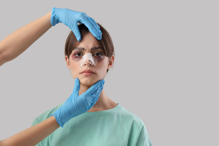 Surgeon hands touching woman's bandaged nose after plastic surgery on gray backgroundの写真素材