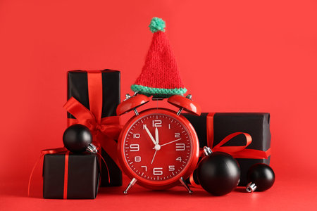 Alarm clock, gift boxes, Christmas balls and elf hat on red background
