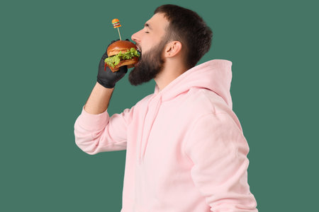 Young man eating tasty burger on green backgroundの写真素材