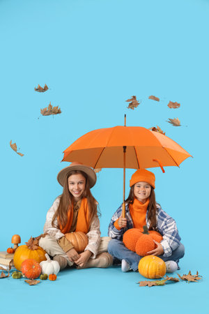 Cute girls in autumn clothes with pumpkins and umbrella sitting on blue backgroundの写真素材