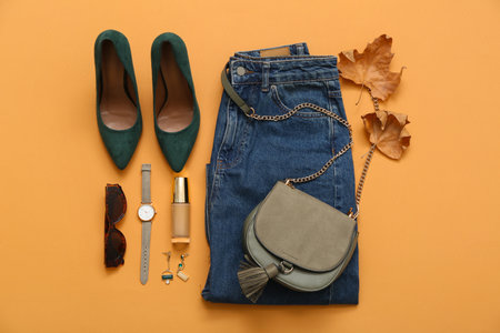 Stylish female bag with jeans, shoes, accessories and autumn leaves on orange backgroundの写真素材