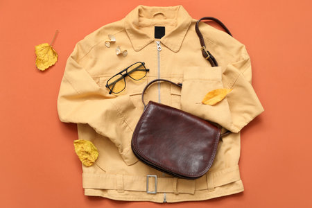 Stylish female bag with jacket, glasses, earrings and autumn leaves on orange backgroundの写真素材