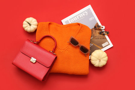 Stylish female bag with knitted sweater, magazine, accessories and autumn decor on red backgroundの写真素材