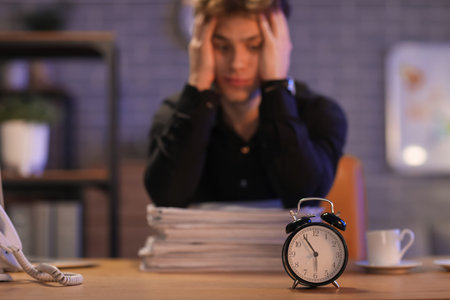 Alarm clock on table against stressed businessman trying to meet deadline in office at night,...