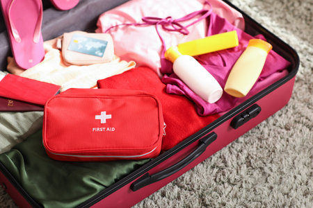 Suitcases with first aid kit, stylish clothes and bottles of sunscreen cream in roomの写真素材